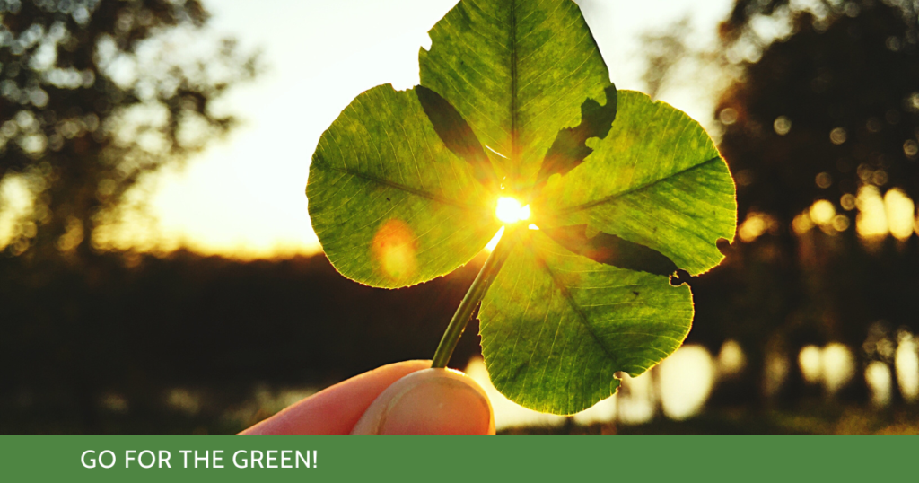 A hand holding a lucky four-leaf clover up to the horizon and the sunshine peaks through a small opening in the center of the clover where all four leaves meet.