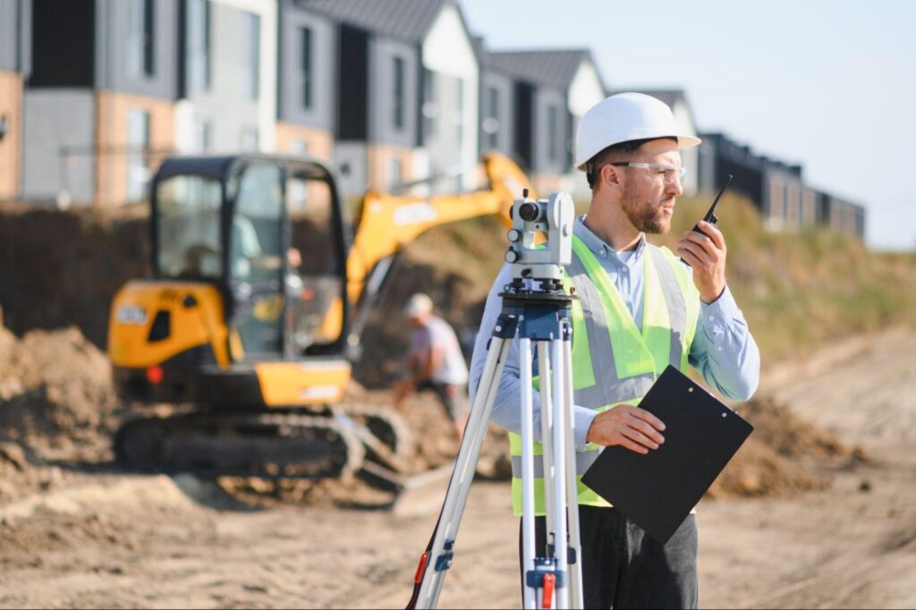 A male land surveyor wearing a white hard hat, safety glasses, and a yellow high-visibility vest stands on a dirt construction site. He is holding a black clipboard and speaking into a two-way radio, with a total station surveying instrument set up on a tripod in front of him. In a blurred background, a yellow mini-excavator and newly constructed townhomes are visible, demonstrating the active field work of engineering consultants.