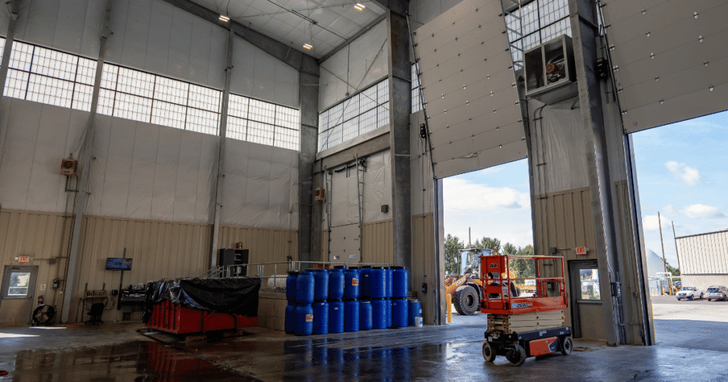 An industrial waste processing warehouse with blue storage barrels, a red dumpster, and a scissor lift, visually illustrating what is biogas and how it is managed.